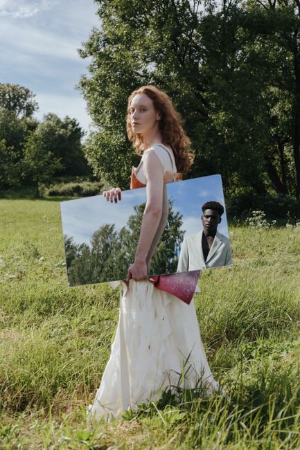 Woman holding a mirror in a field, reflecting a man in a suit, blending nature and art concepts.