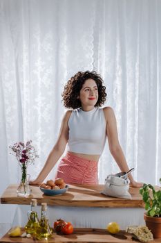 A cheerful woman with curly hair preparing food in a sunlit kitchen.