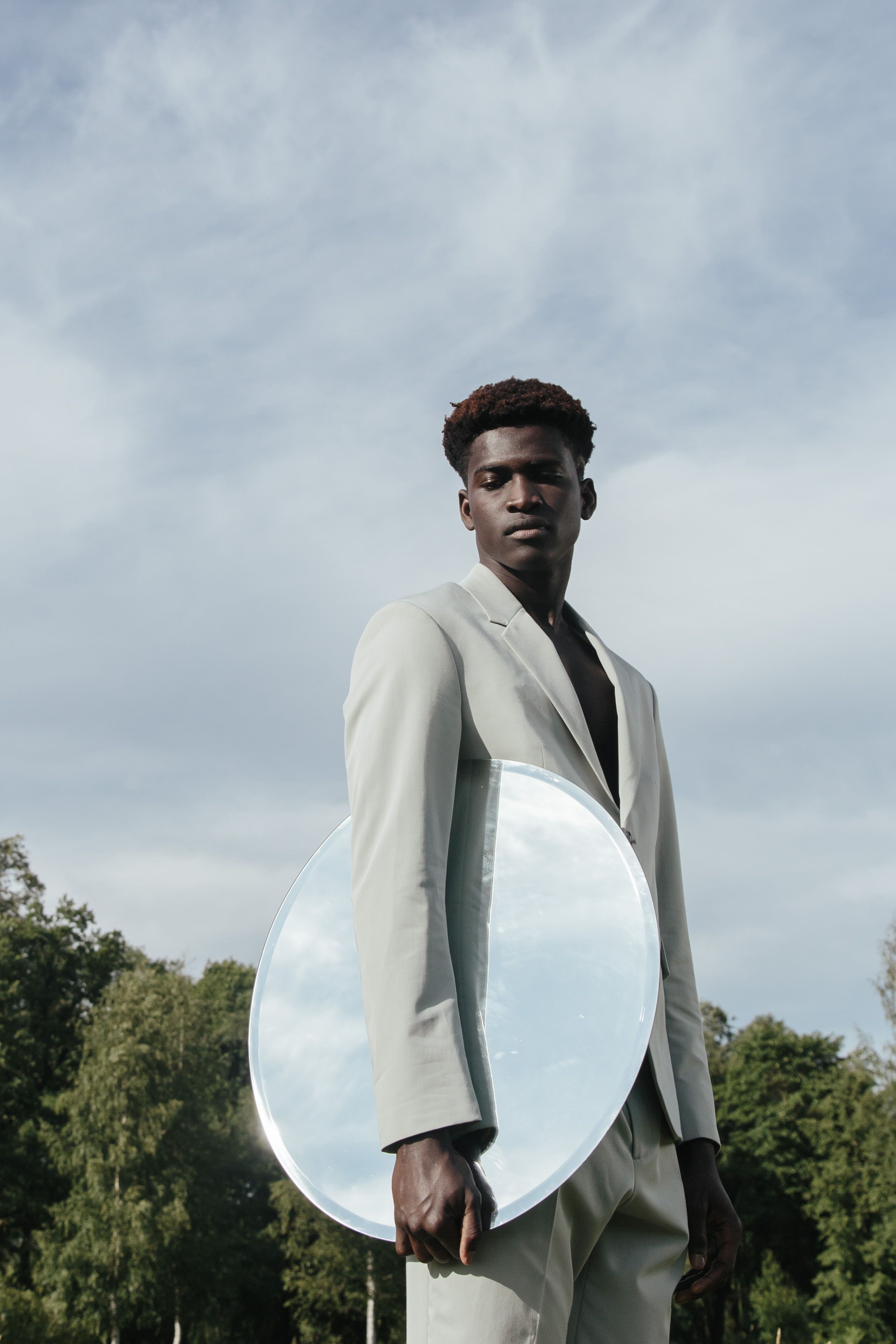 African American man in a light suit holding a round mirror outdoors, reflecting the sky and trees.