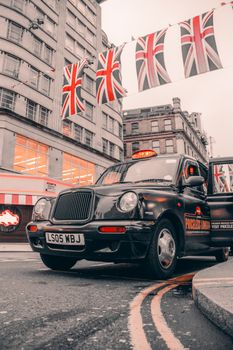 Black London taxi cab under Union Jack flags, showcasing UK city life.