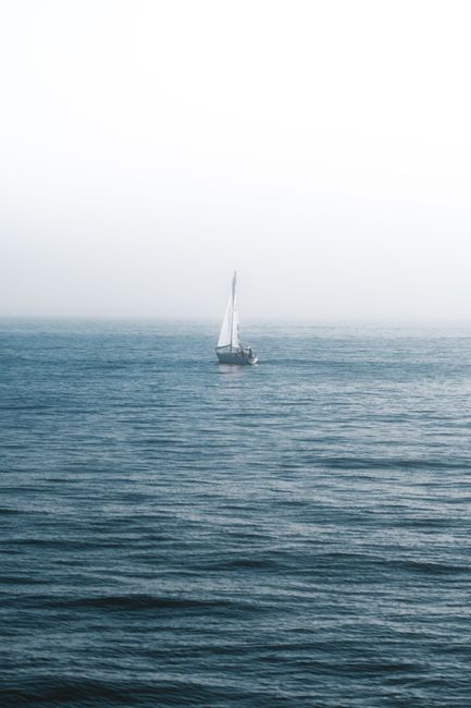 A lone sailboat glides across a calm, foggy ocean under a bright sky.