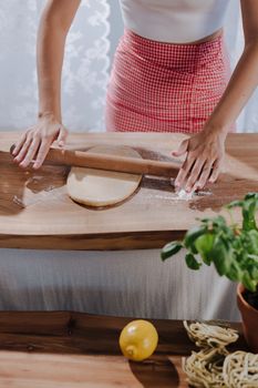 Woman rolling dough on wooden table with lemon and pasta nearby indoors.