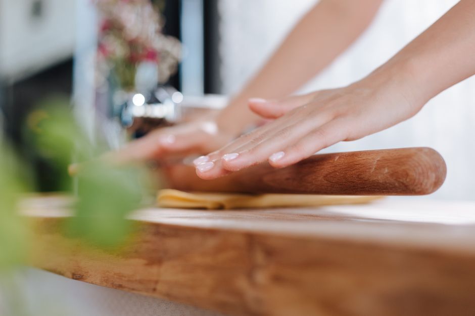 A mound of yellow pasta dough being kneaded by hand on a floured wooden board, with a bench scraper and cracked eggshells nearby.
