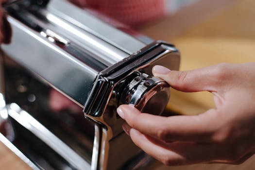 Close-up view of hands adjusting a shiny metal pasta maker for Italian cuisine preparation.
