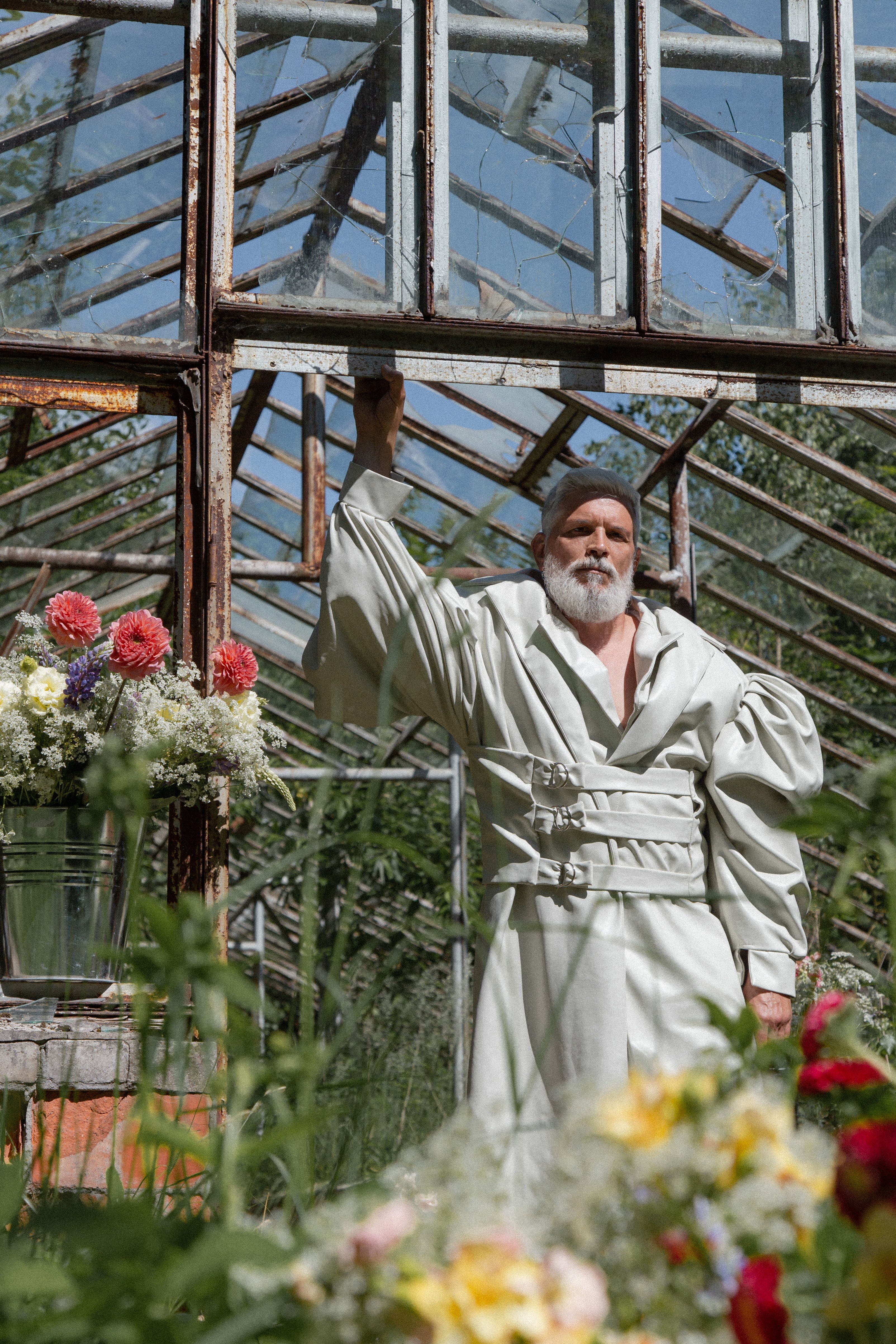 Senior man in stylish fashion coat posing in a rustic greenhouse with colorful flowers.