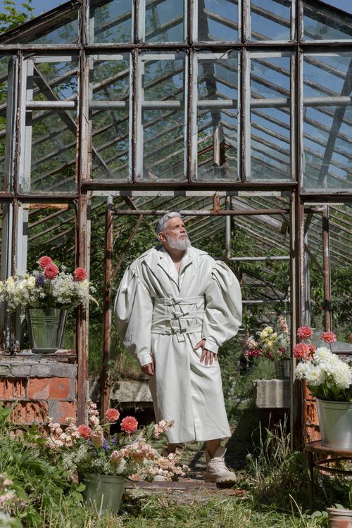 Free Man in stylish attire posing in an abandoned, rustic greenhouse surrounded by vibrant flowers. Stock Photo
