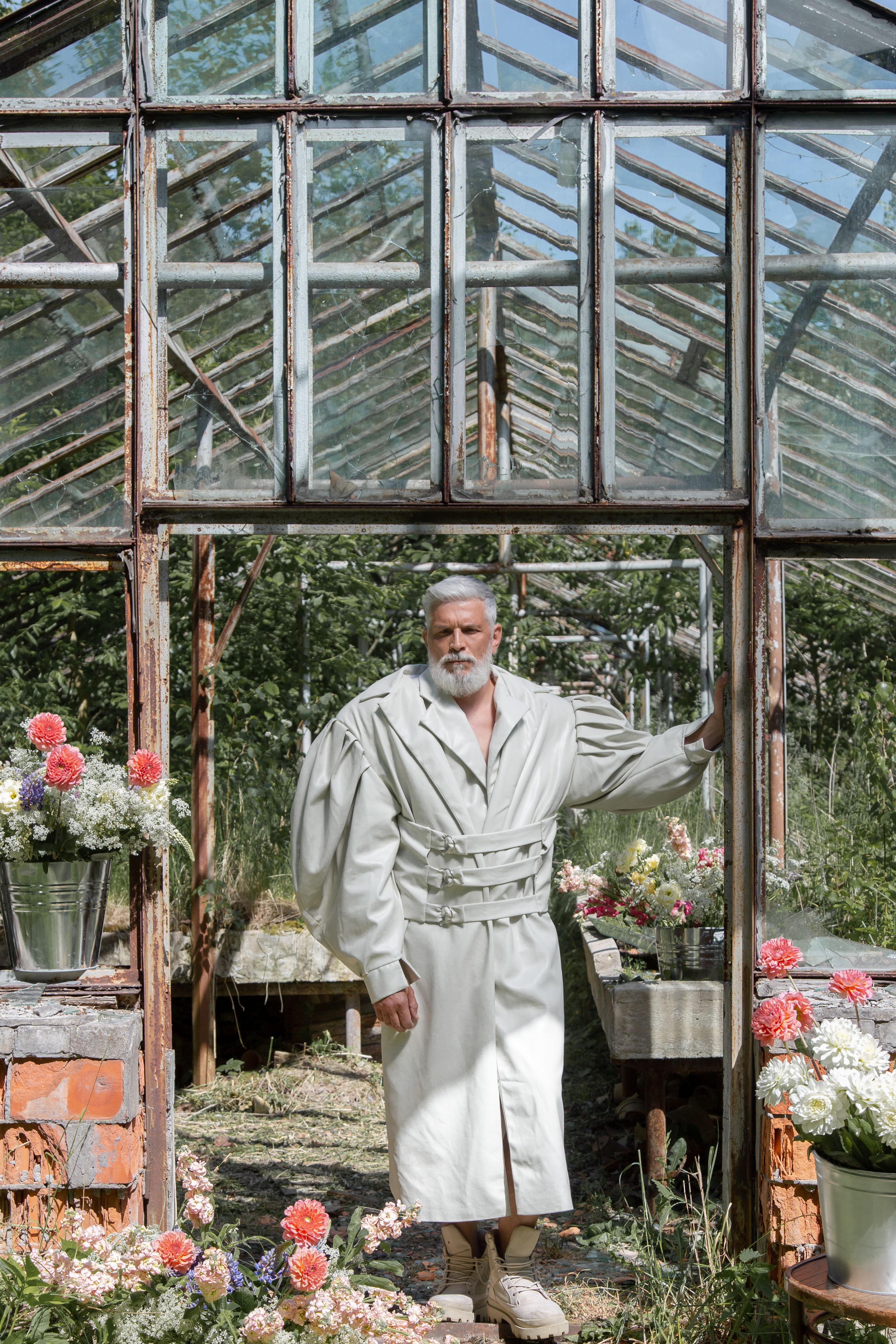 Elderly man in unique trench coat inside an old greenhouse surrounded by flowers.