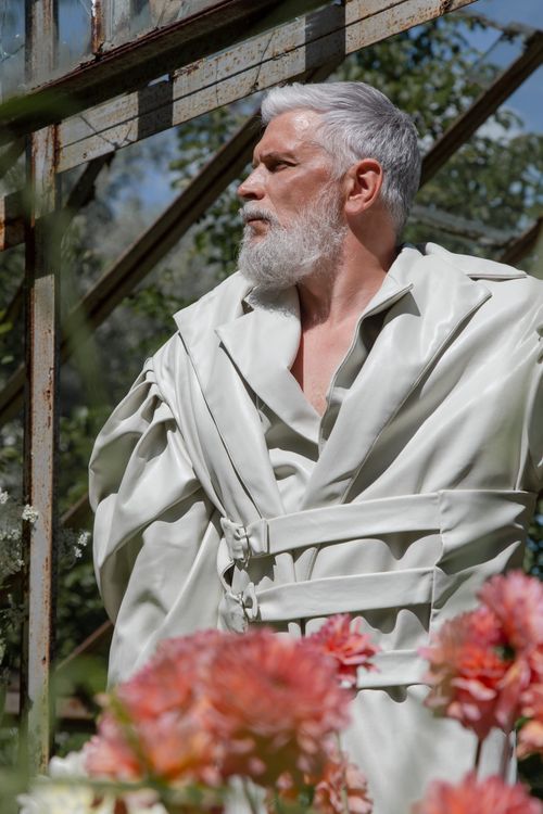 Free Elderly man poses in a stylish trench coat outdoors with flowers in the foreground. Stock Photo