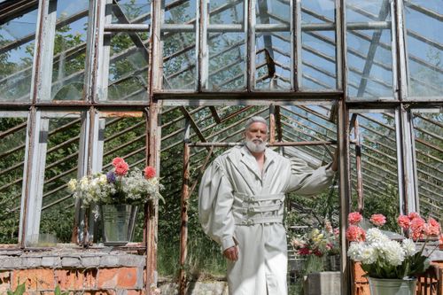 Free Senior man in white coat standing in an abandoned greenhouse surrounded by flowers. A blend of nature and rustic architecture. Stock Photo