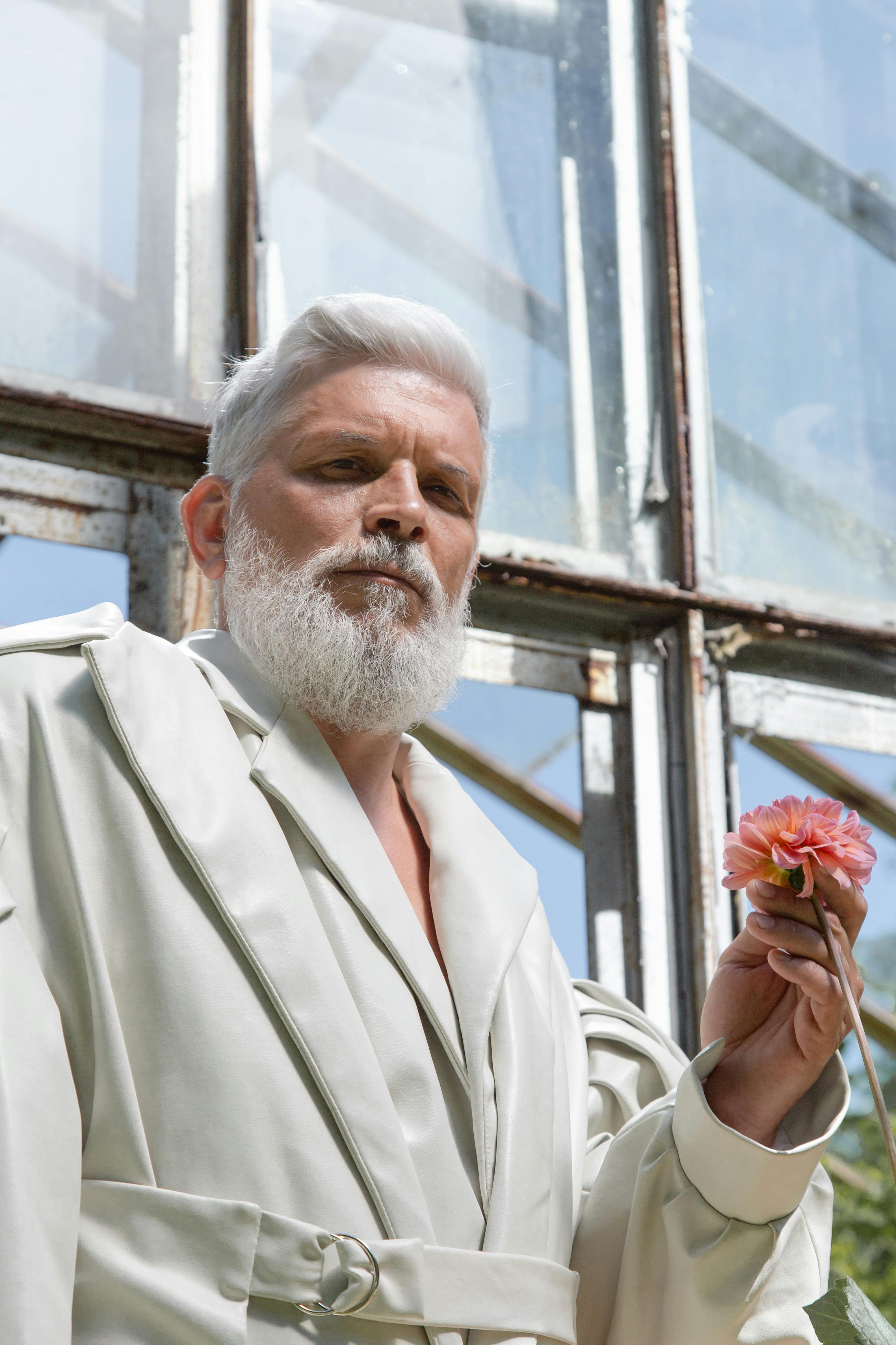 Sophisticated senior man in white robe holding a pink flower in a sunlit room.