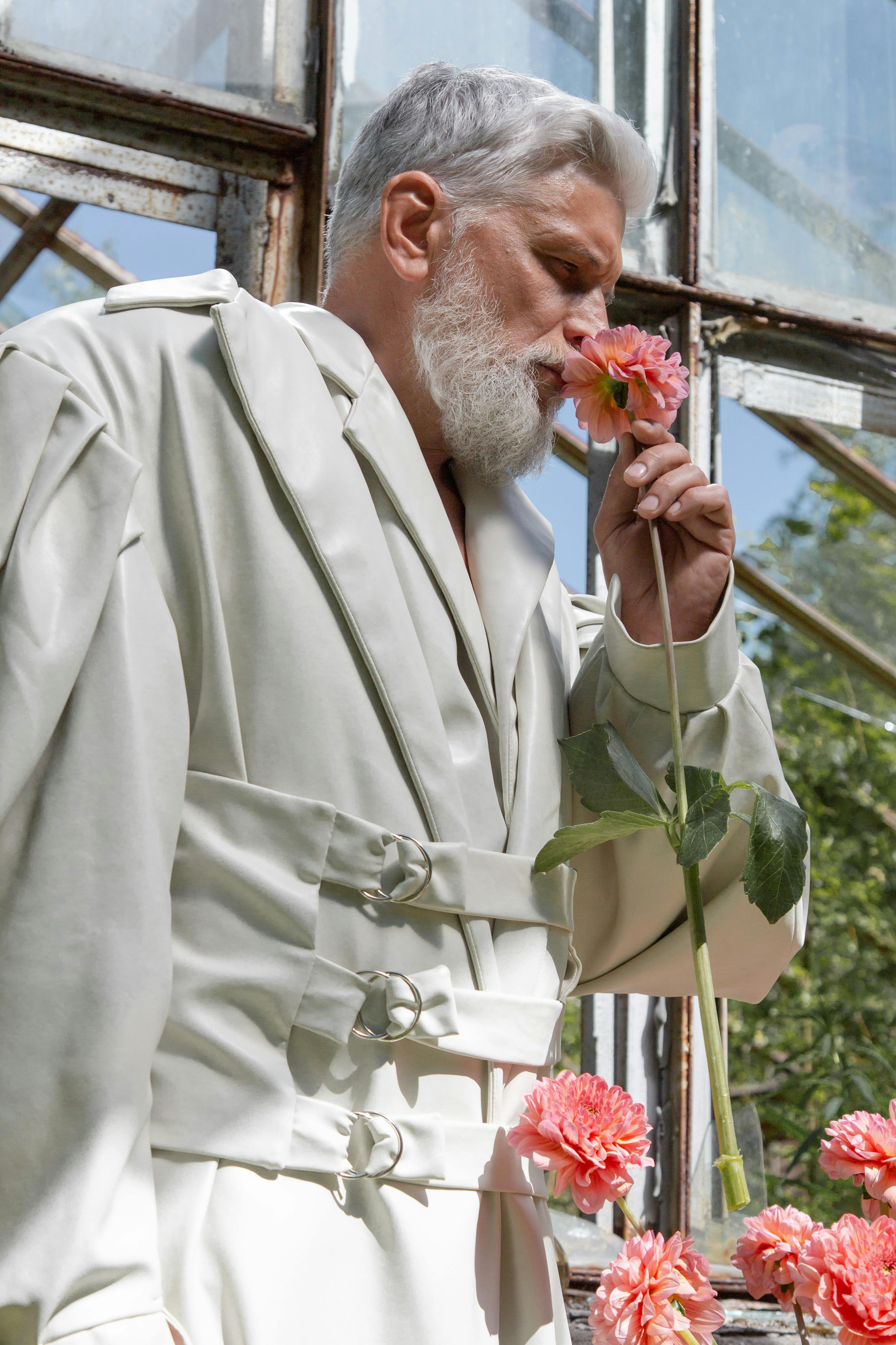 A senior man in a stylish coat enjoys the fragrance of a flower in a greenhouse setting.