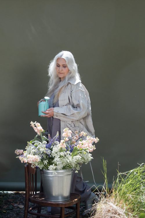 Free Elderly woman holding a watering can beside a floral arrangement in a garden on a sunny day. Stock Photo