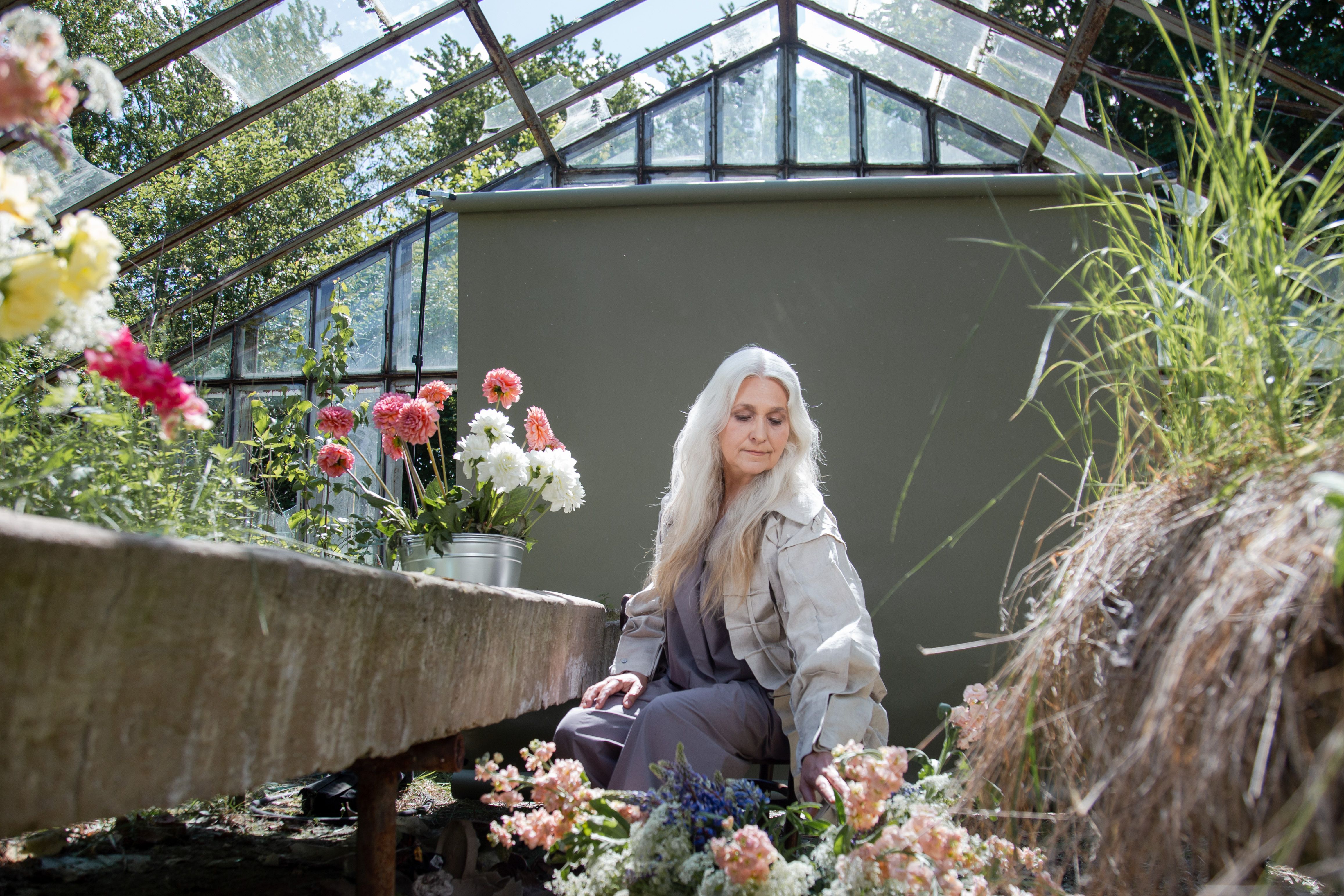 Mature woman arranging flowers inside a sunlit greenhouse.