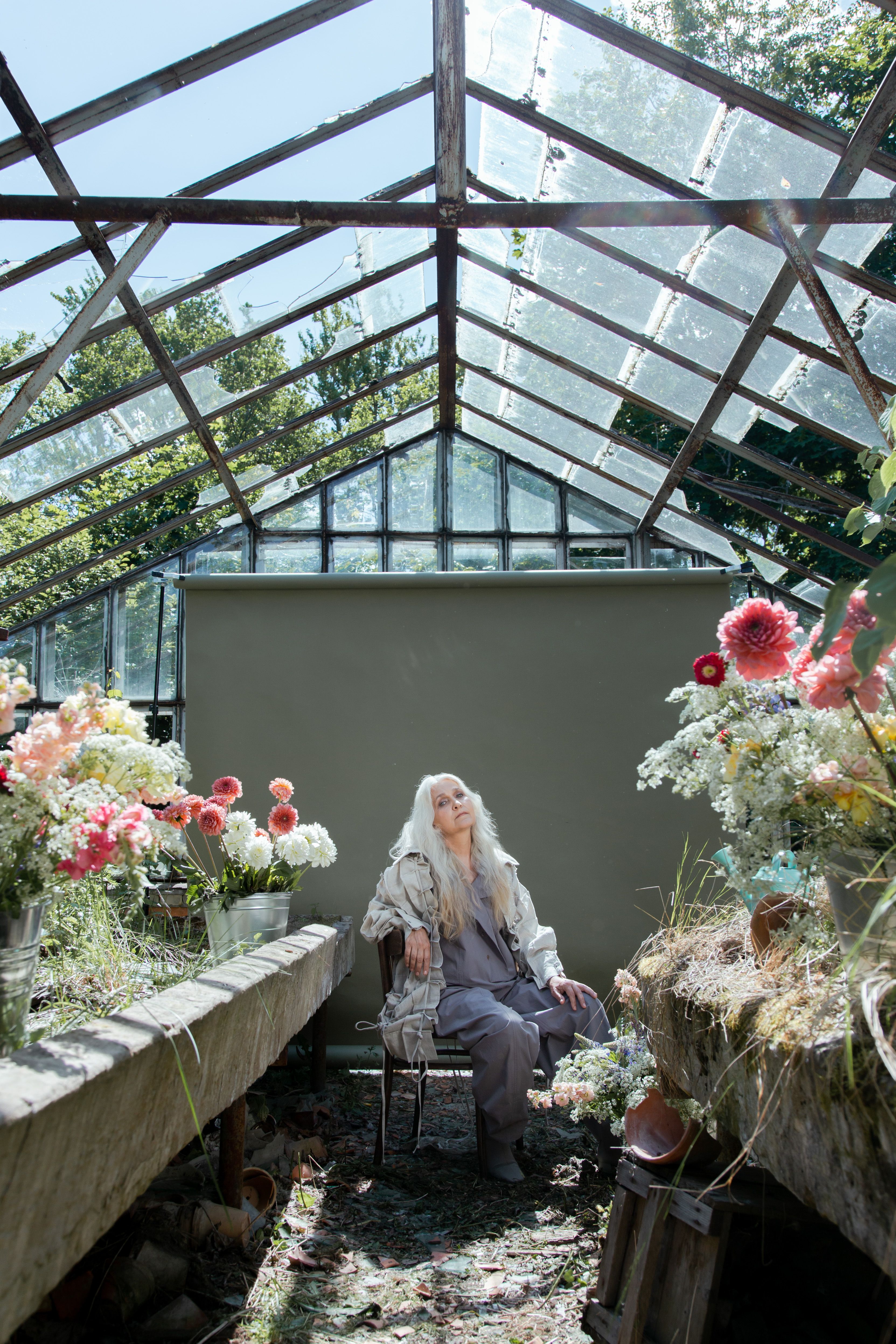 A senior woman with long gray hair sits elegantly in a flower-filled greenhouse, basking in sunlight.