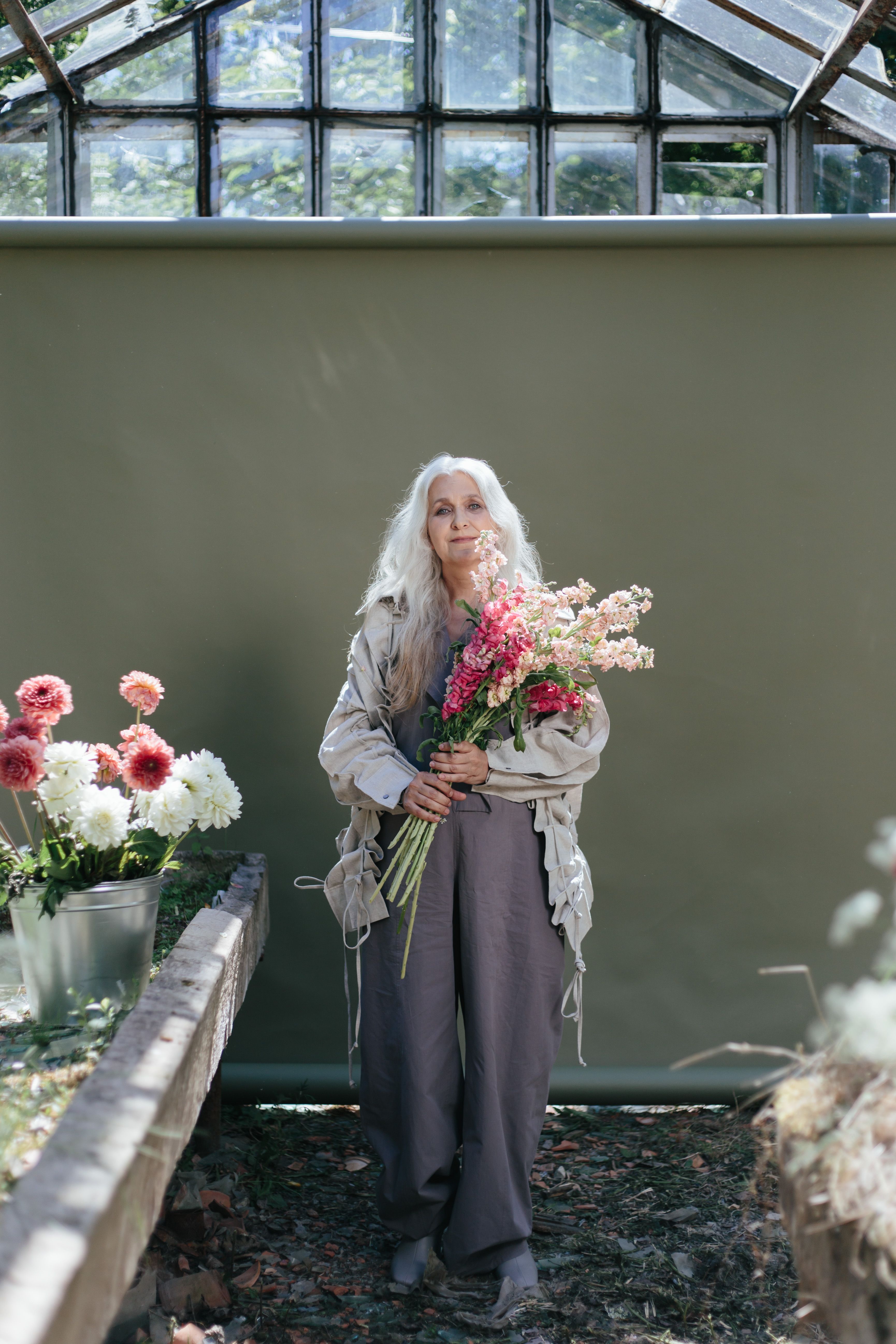 Senior woman with long hair holding a vibrant bouquet in an elegant greenhouse setting.