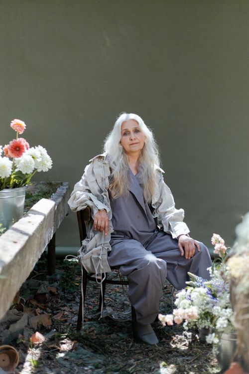 Free Senior woman in gray sitting indoors surrounded by flowers, creating a serene scene. Stock Photo