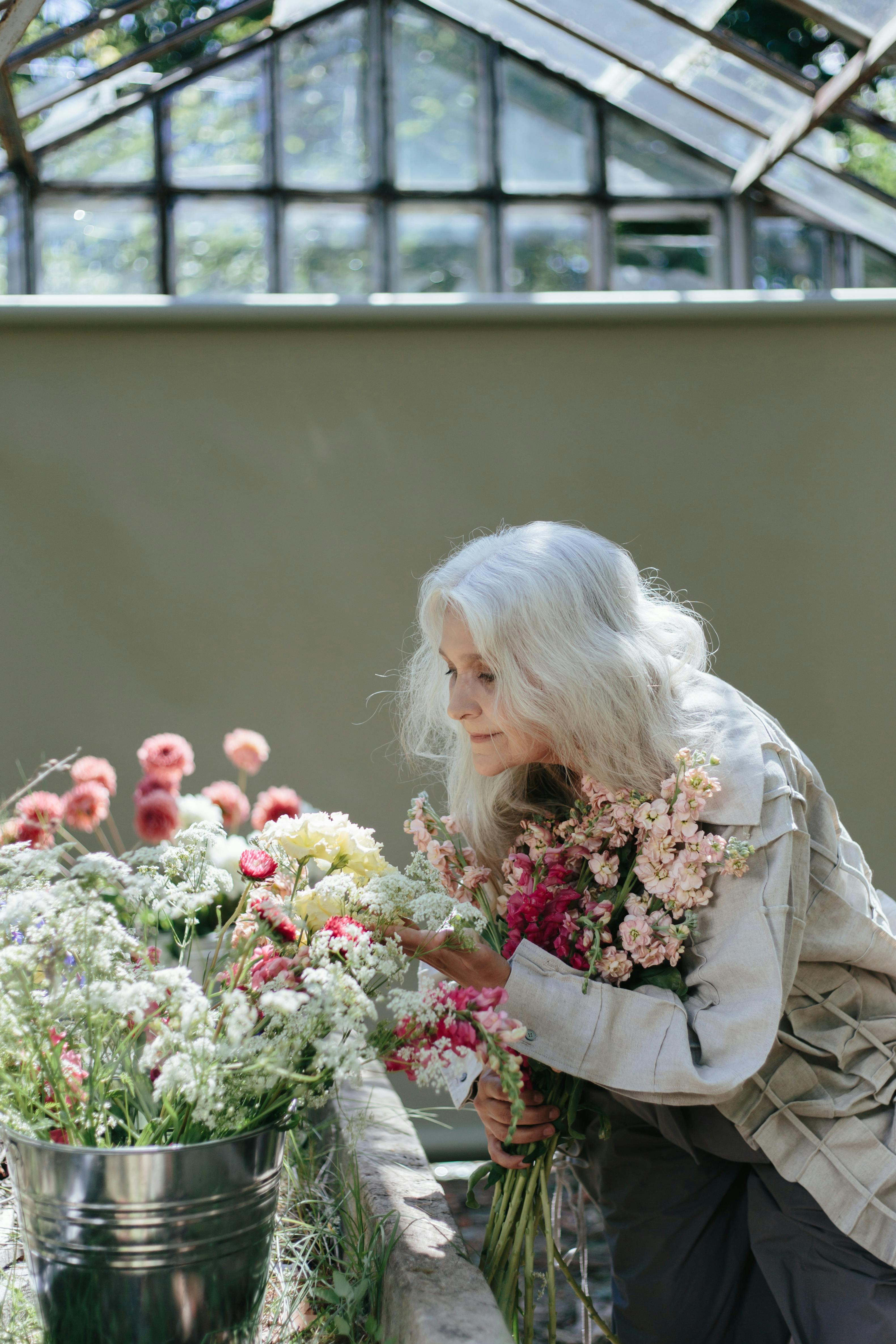 Elderly woman with gray hair admiring flowers in a greenhouse, exuding tranquility.