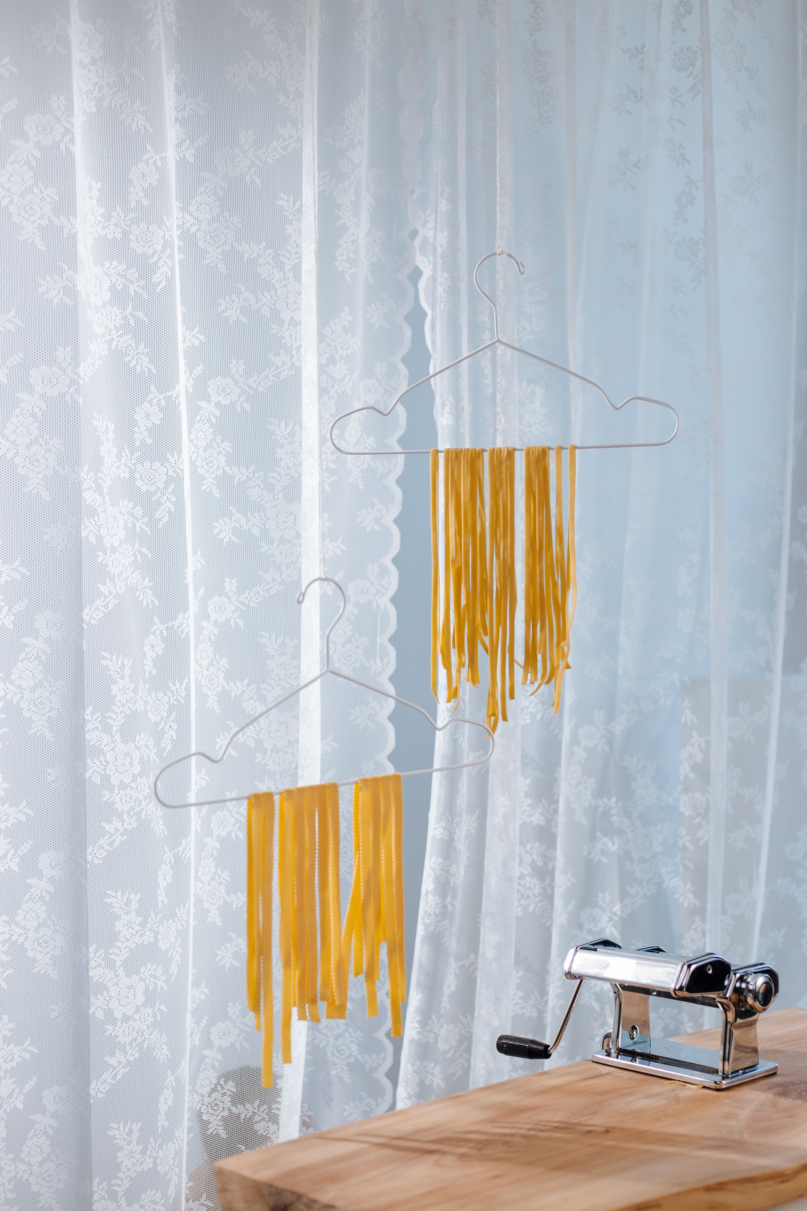 Fresh homemade pasta strands drying on hangers with a pasta machine nearby, creating a rustic kitchen scene.