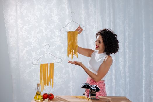Woman drying homemade pasta on hangers with ingredients and pasta machine on table.