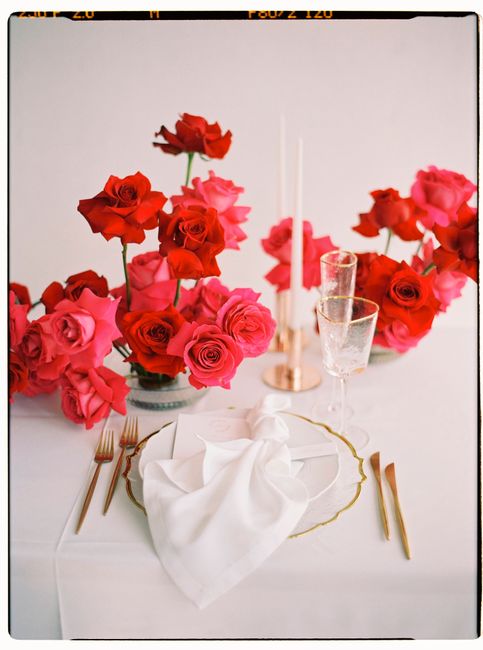 Luxurious table arrangement with red and pink roses, gold cutlery, and candlelight against a white background.