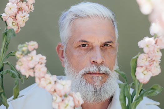 Elderly man with gray hair and beard surrounded by flowers outdoors.