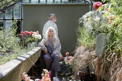 A Elderly Couple Sitting and Standing Near Flowers
