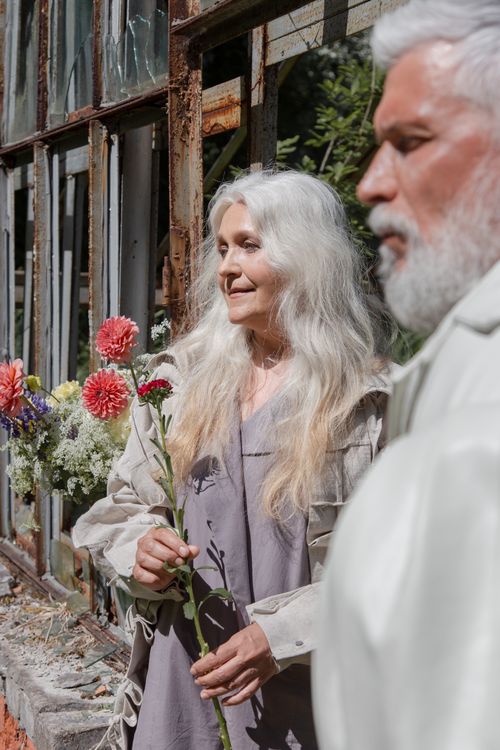 Free Elderly couple enjoying the outdoors, holding flowers beside a rustic window. Stock Photo