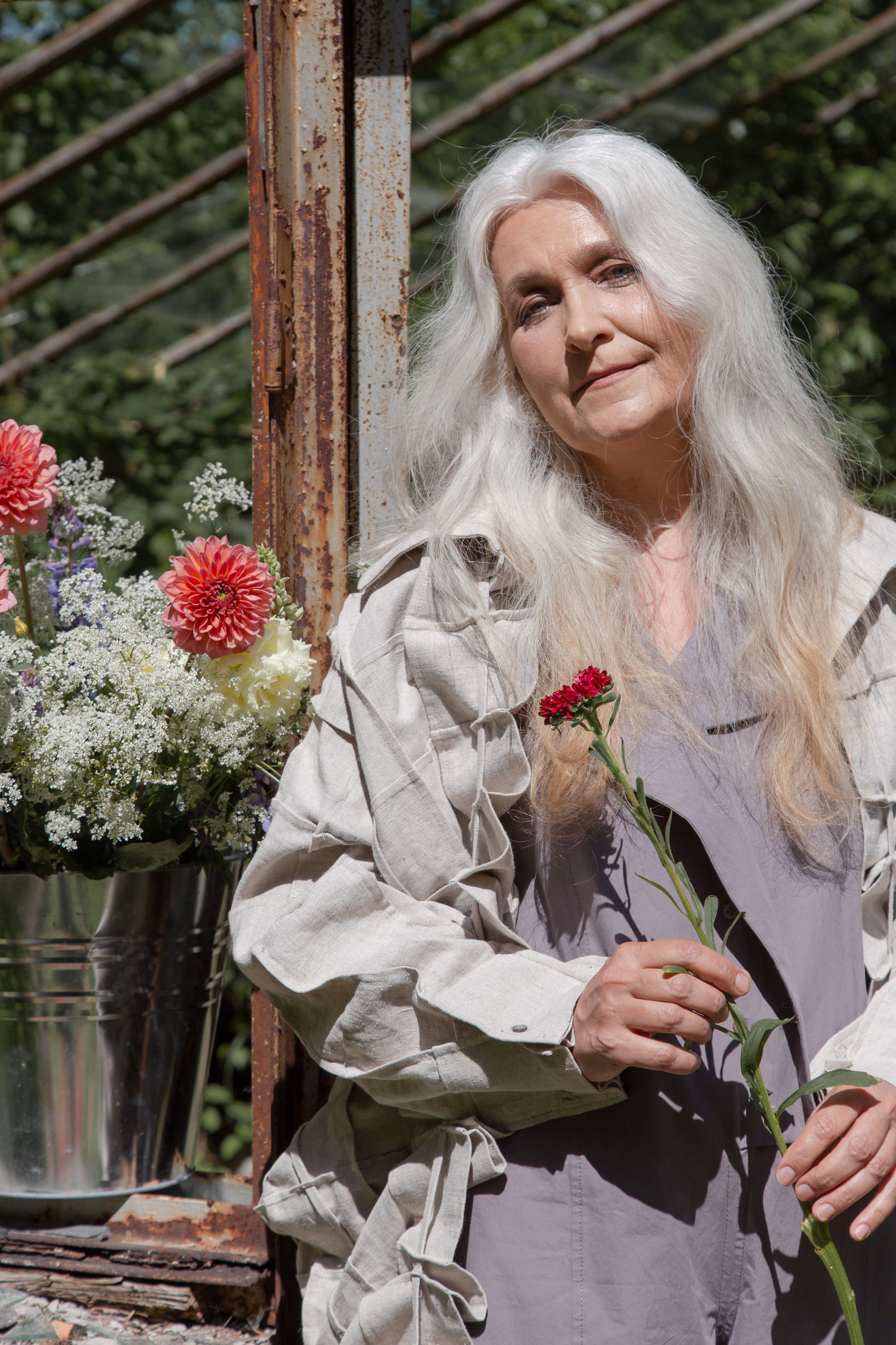 Serene senior woman in a garden setting, holding a red flower with vibrant blooms nearby.