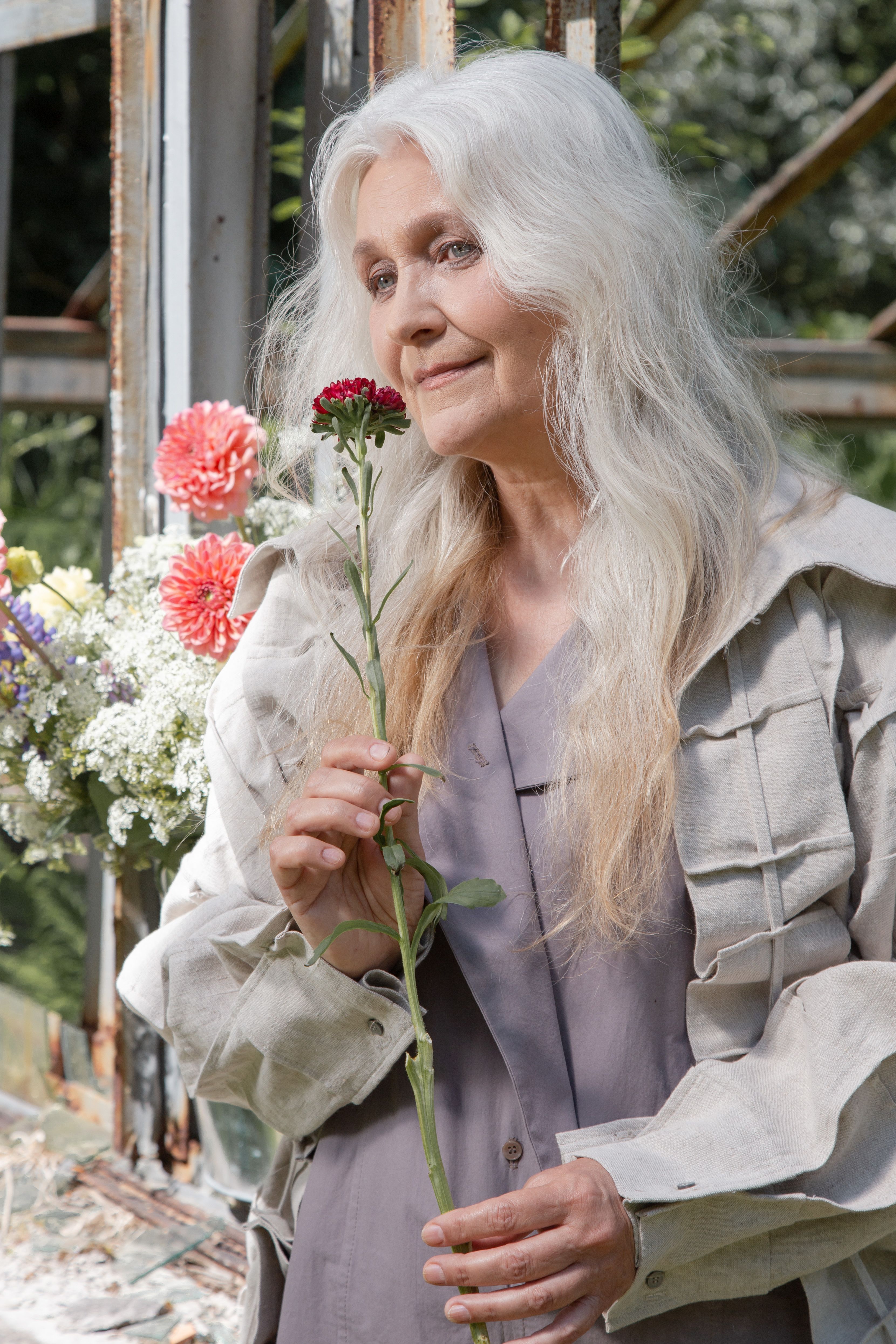 An elderly woman with long gray hair holding a red carnation, surrounded by flowers in a rustic outdoor setting.