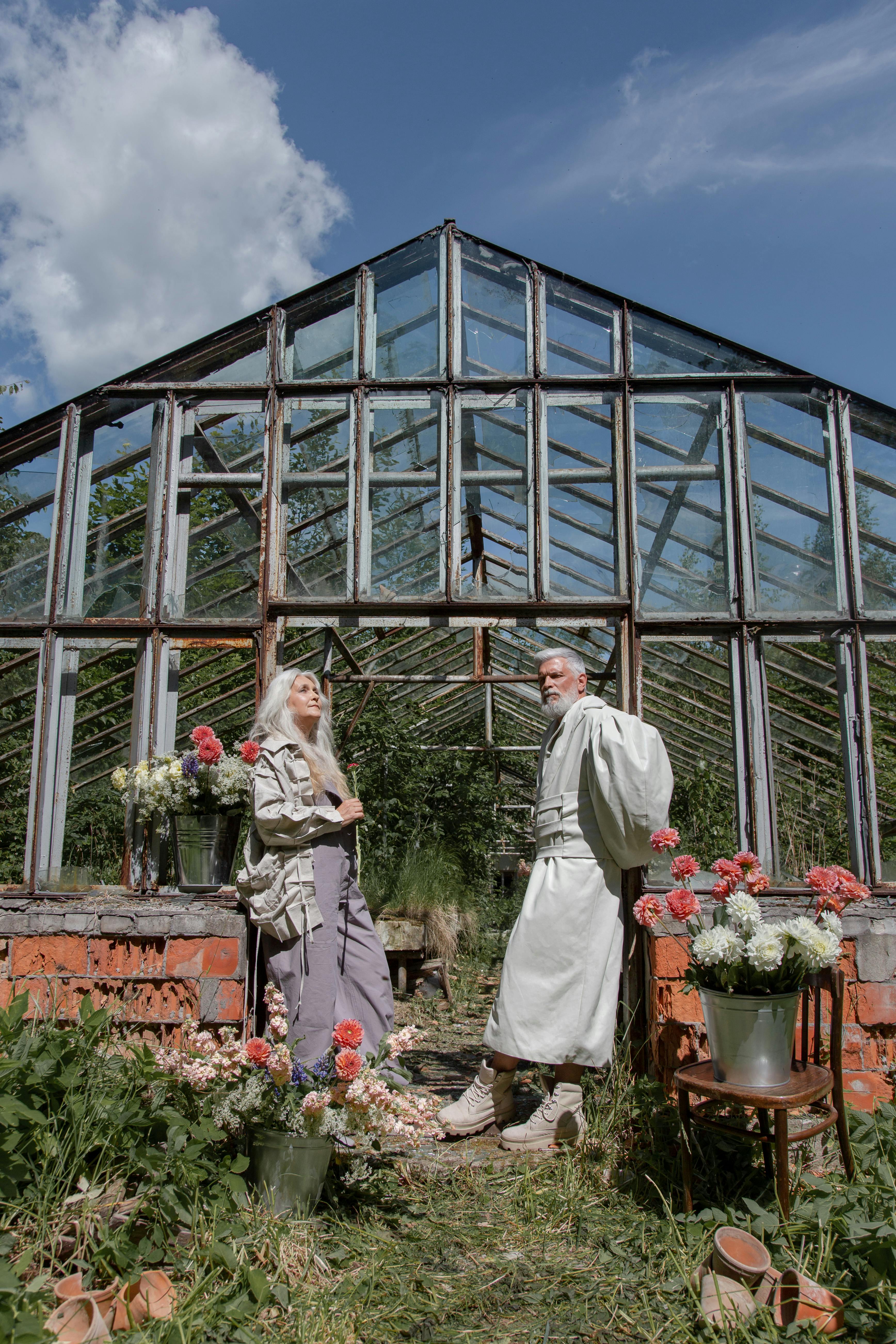 An elderly couple spending time together outside a rustic greenhouse surrounded by flowers.