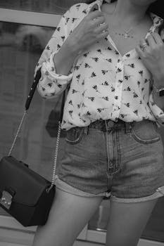 Black and white photo of a woman in shorts and patterned shirt holding a handbag.
