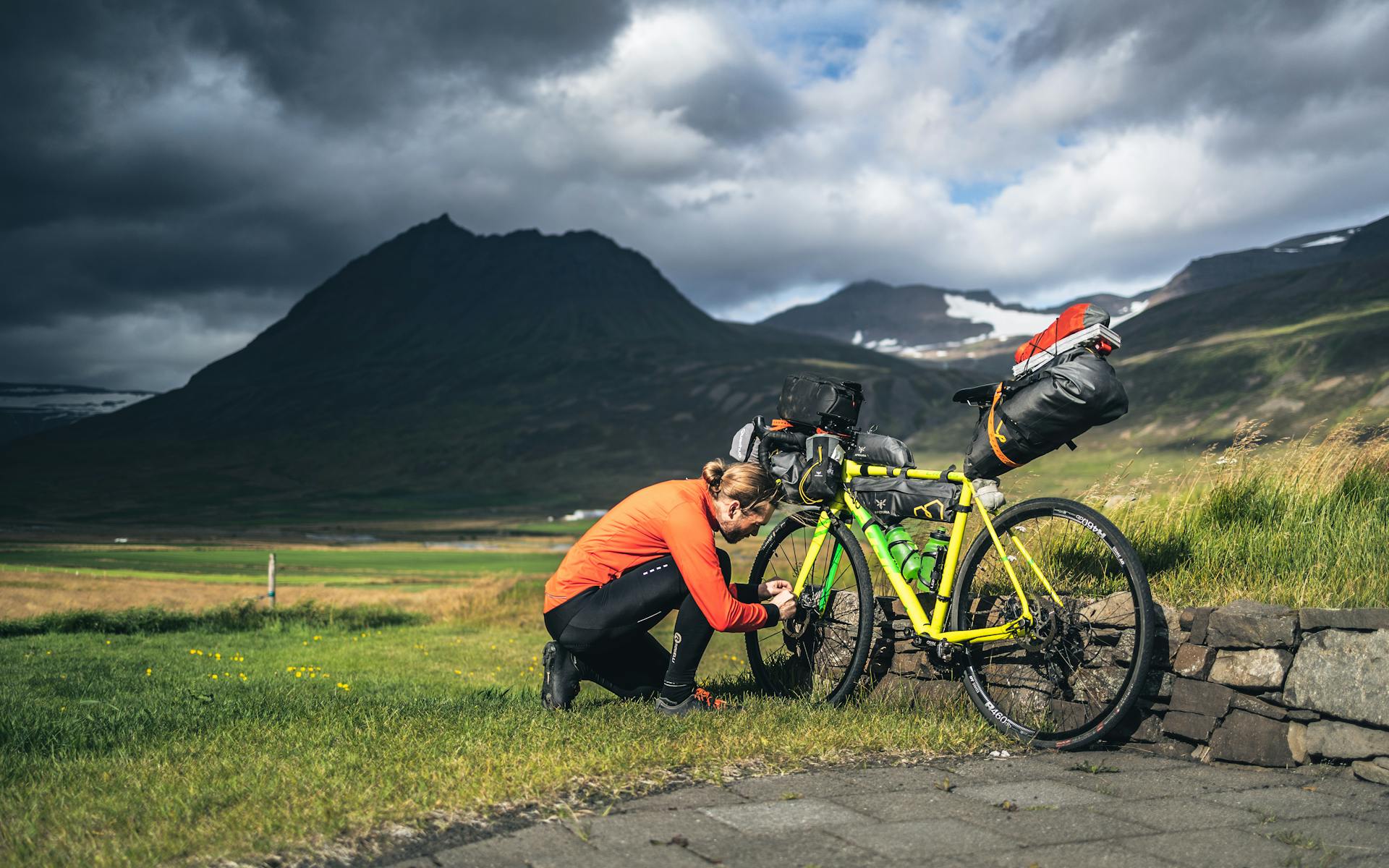 Cyclist adjusting gear on loaded bike in Iceland's rugged landscape under dramatic skies.