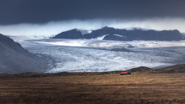 A breathtaking Icelandic landscape featuring a glacier, mountains, and a small red-roofed hut under a dramatic sky.