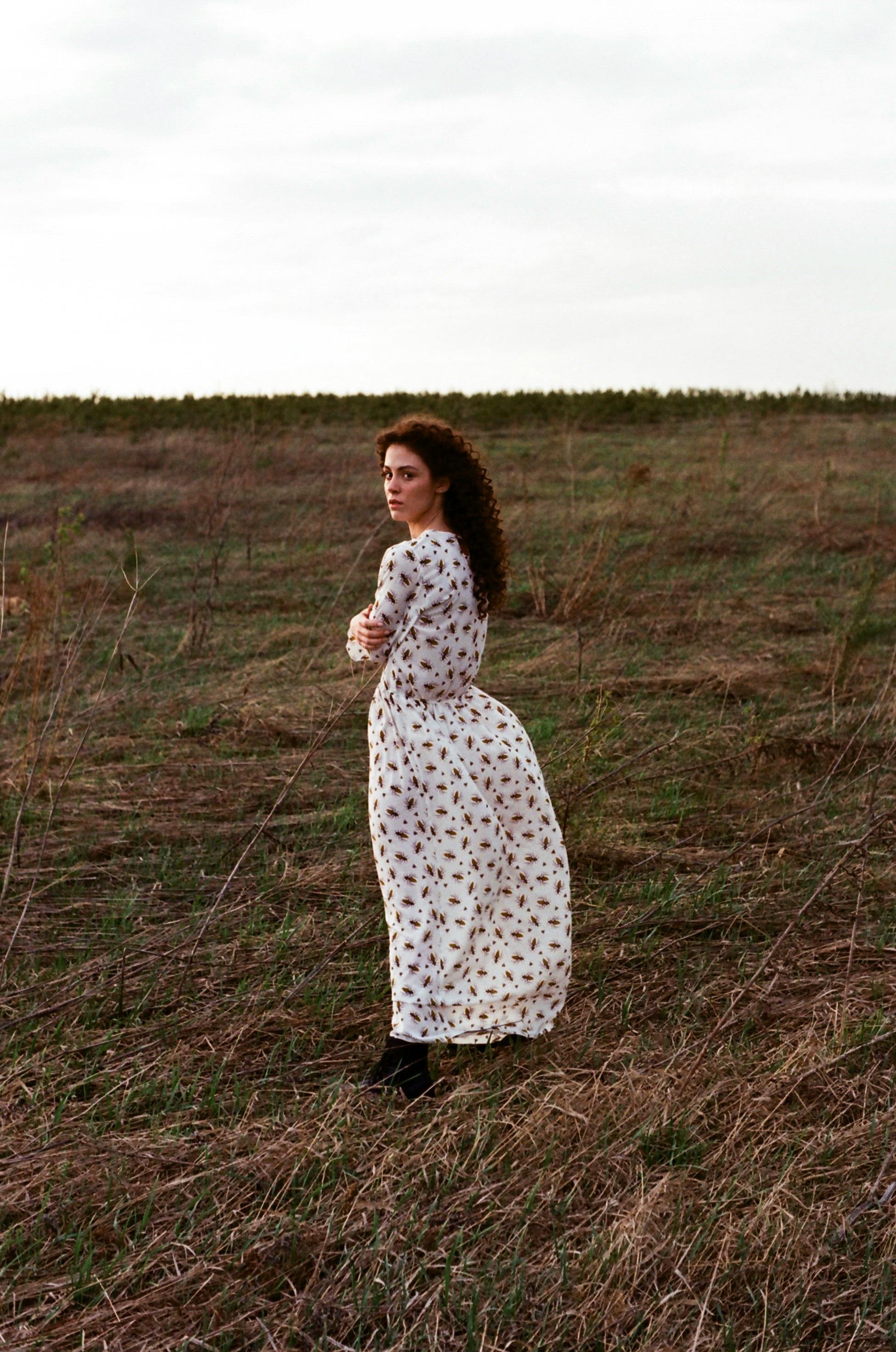 A woman in a floral dress stands in a rural field, gazing back.