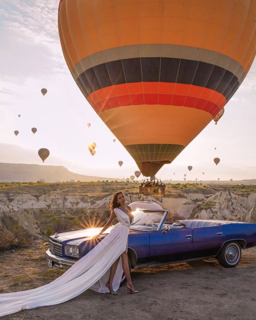 Free A woman in a white dress beside a classic blue convertible at sunrise with hot air balloons. Stock Photo