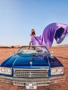 Woman in elegant purple dress standing on a vintage blue car in a desert landscape.