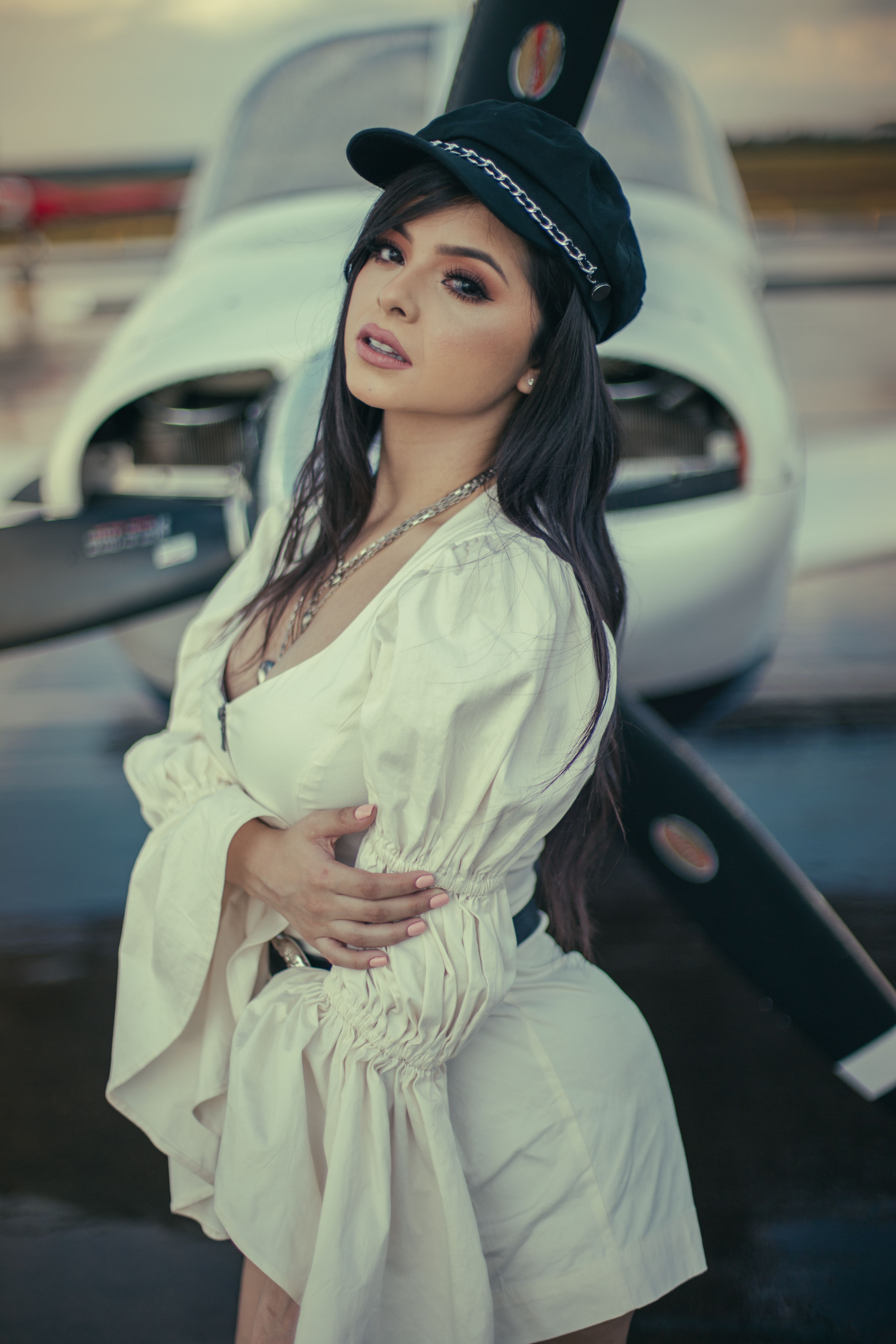 Chic outdoor portrait of a woman in fashion attire standing beside a private airplane with a confident pose.