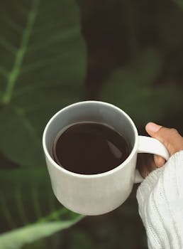 A cozy close-up of a hand holding a coffee mug against lush green foliage.