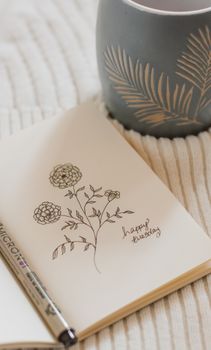 A delicate floral sketch in a notebook beside a stylized mug on a textile surface, inspiring creativity.