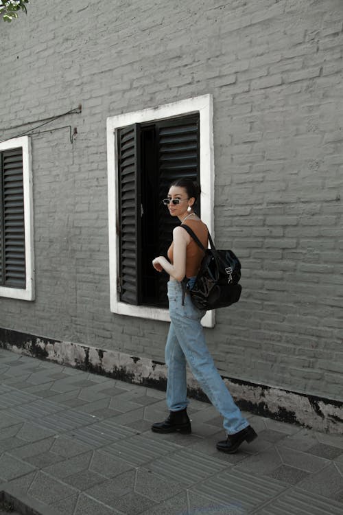 Free Woman in Sleeveless Top and Blue Denim Jeans Walking Beside Gray Concrete Building Stock Photo