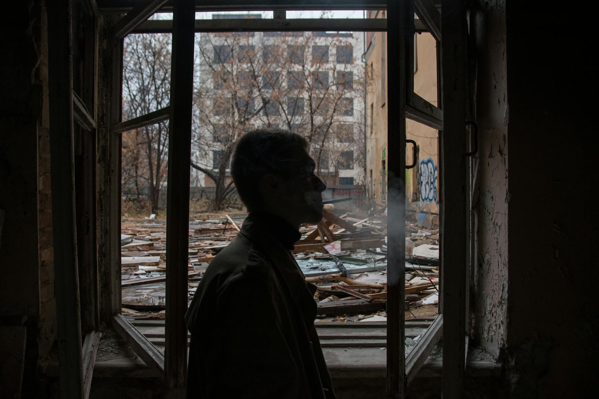 A man smoking inside an abandoned building, surrounded by debris and broken windows.