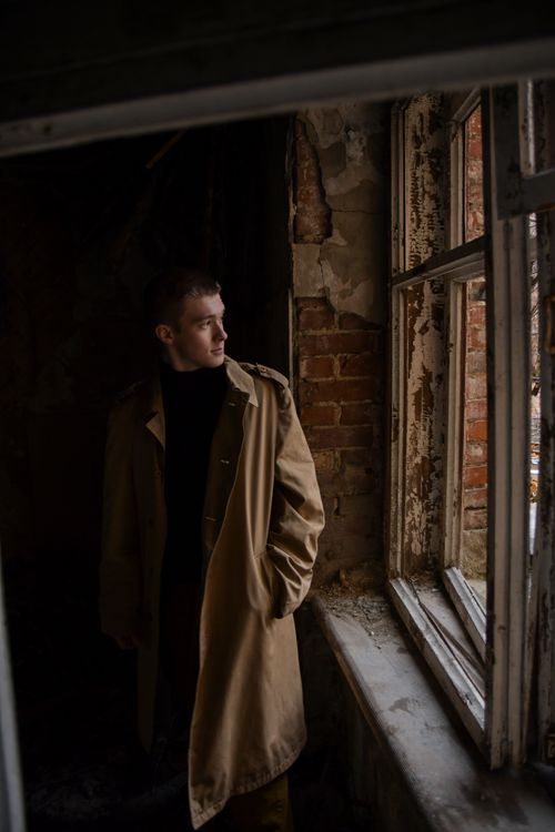 Free A man in a trench coat gazes out a rustic window, creating a mysterious and moody atmosphere. Stock Photo