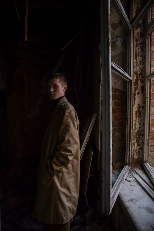 Free A man in a trench coat stands by a window in a dimly lit room, creating a moody atmosphere. Stock Photo