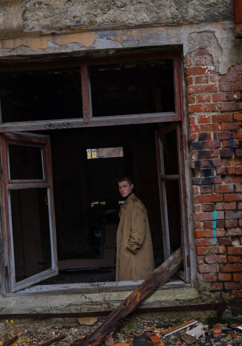 Free A man in a beige coat standing in a broken window of an abandoned and vandalized brick building. Stock Photo