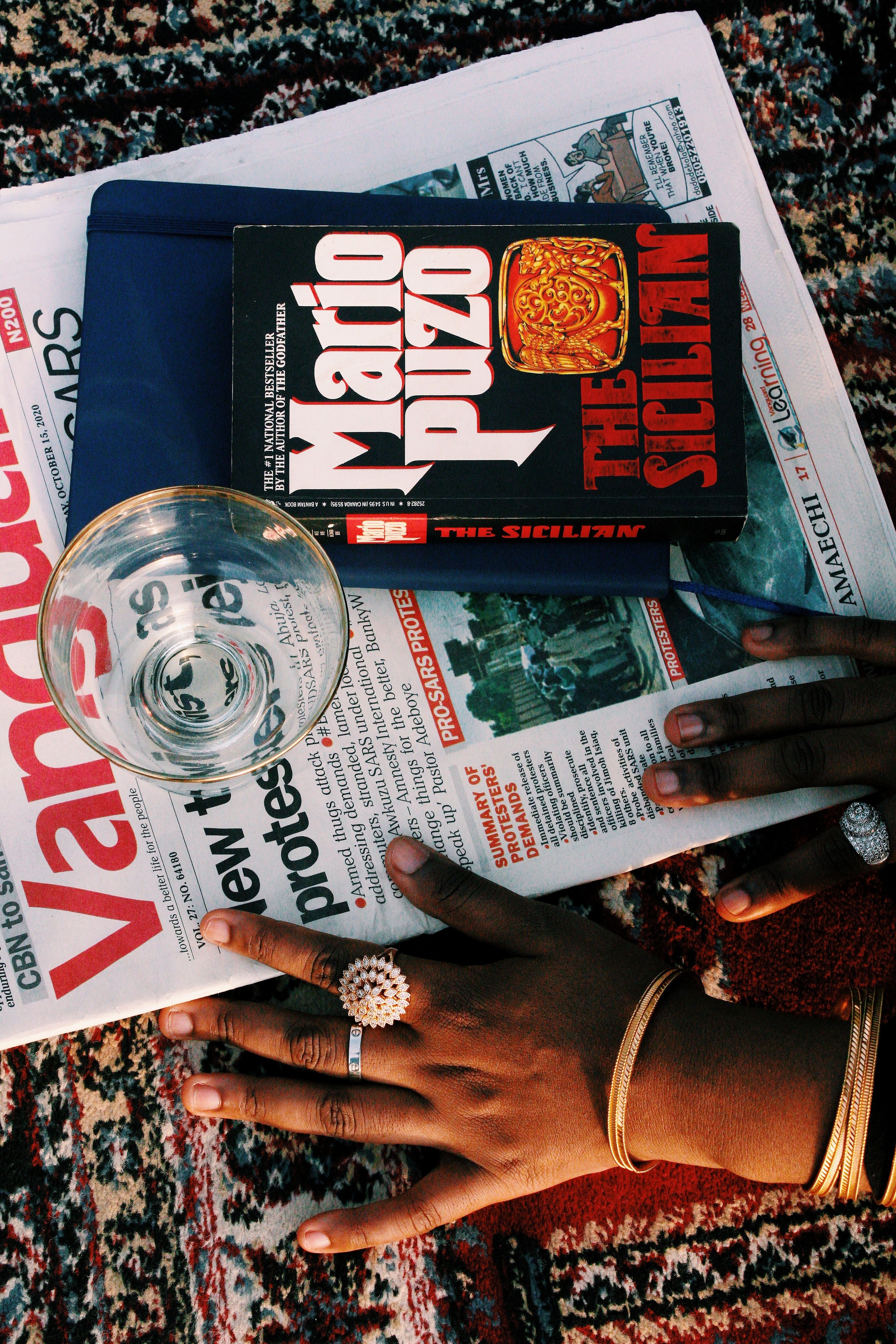 Flatlay of hands with jewelry on book and newspaper on patterned carpet in Nigeria.