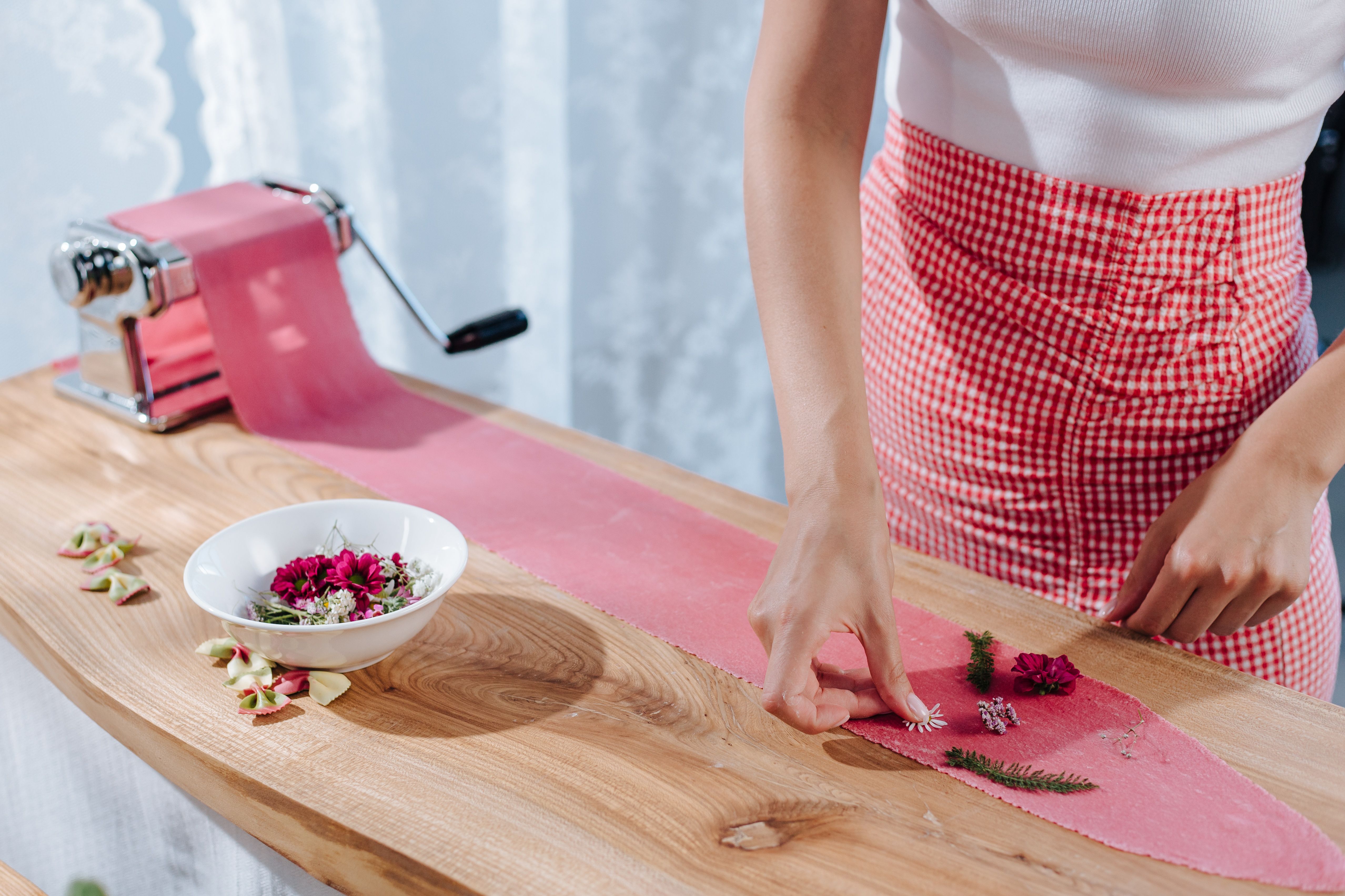 Woman in gingham skirt preparing flower-infused pasta using a pasta maker on a wooden surface.