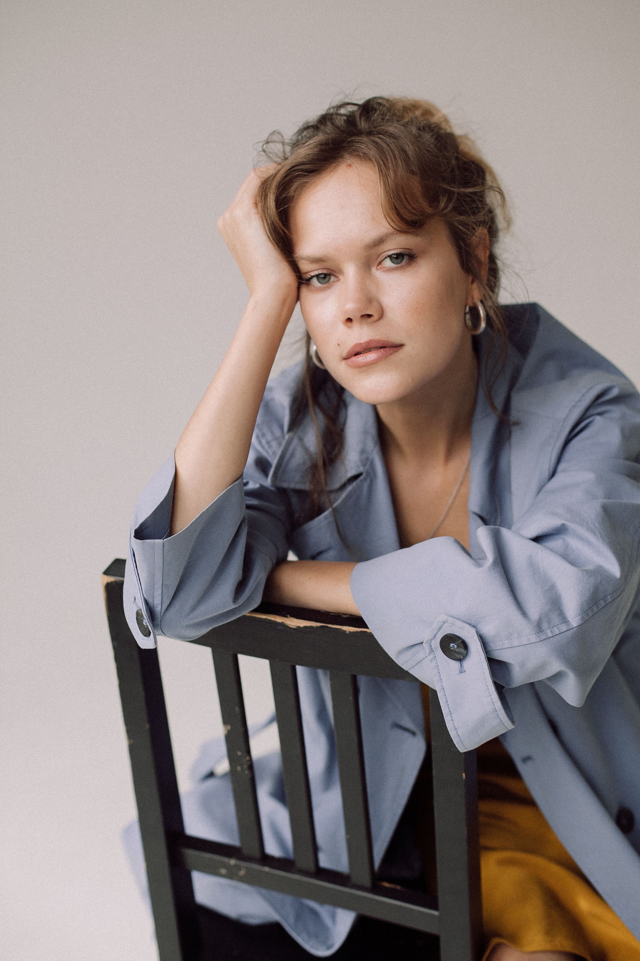 A stylish woman in a blue coat leans on a chair, exuding elegance in a studio portrait.