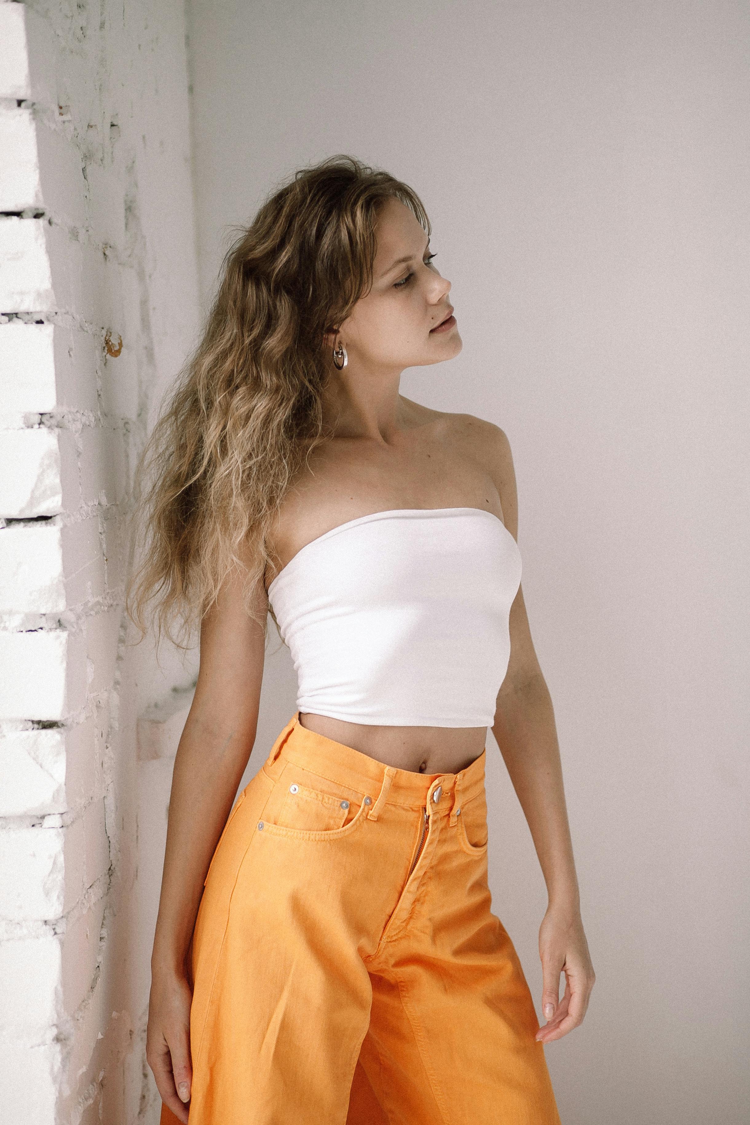 Elegant woman posing in a white tube top and orange pants against a minimalist brick wall indoors.