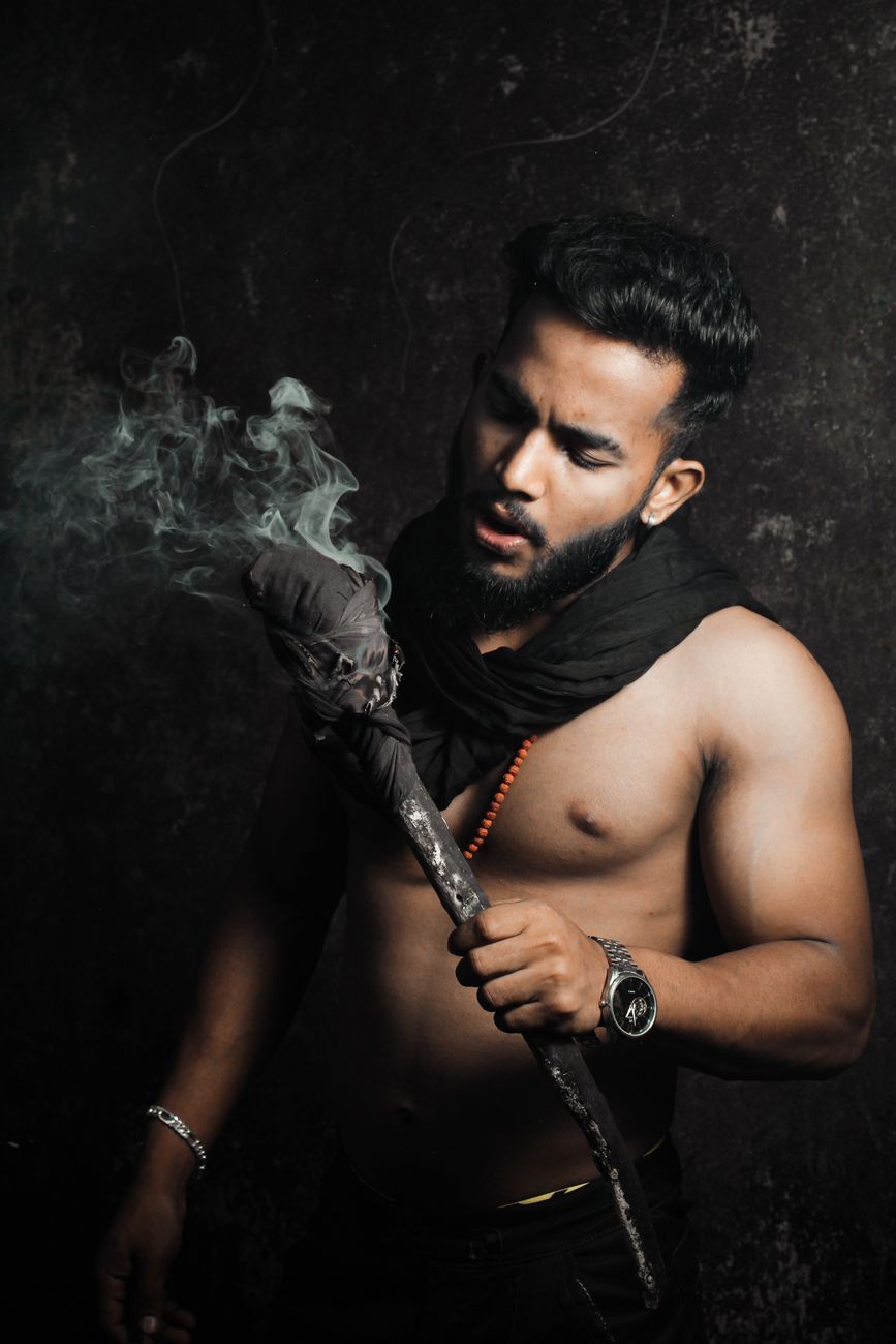 Shirtless man holding a smoking torch in a dramatic studio setting, emphasizing mood and mystery.