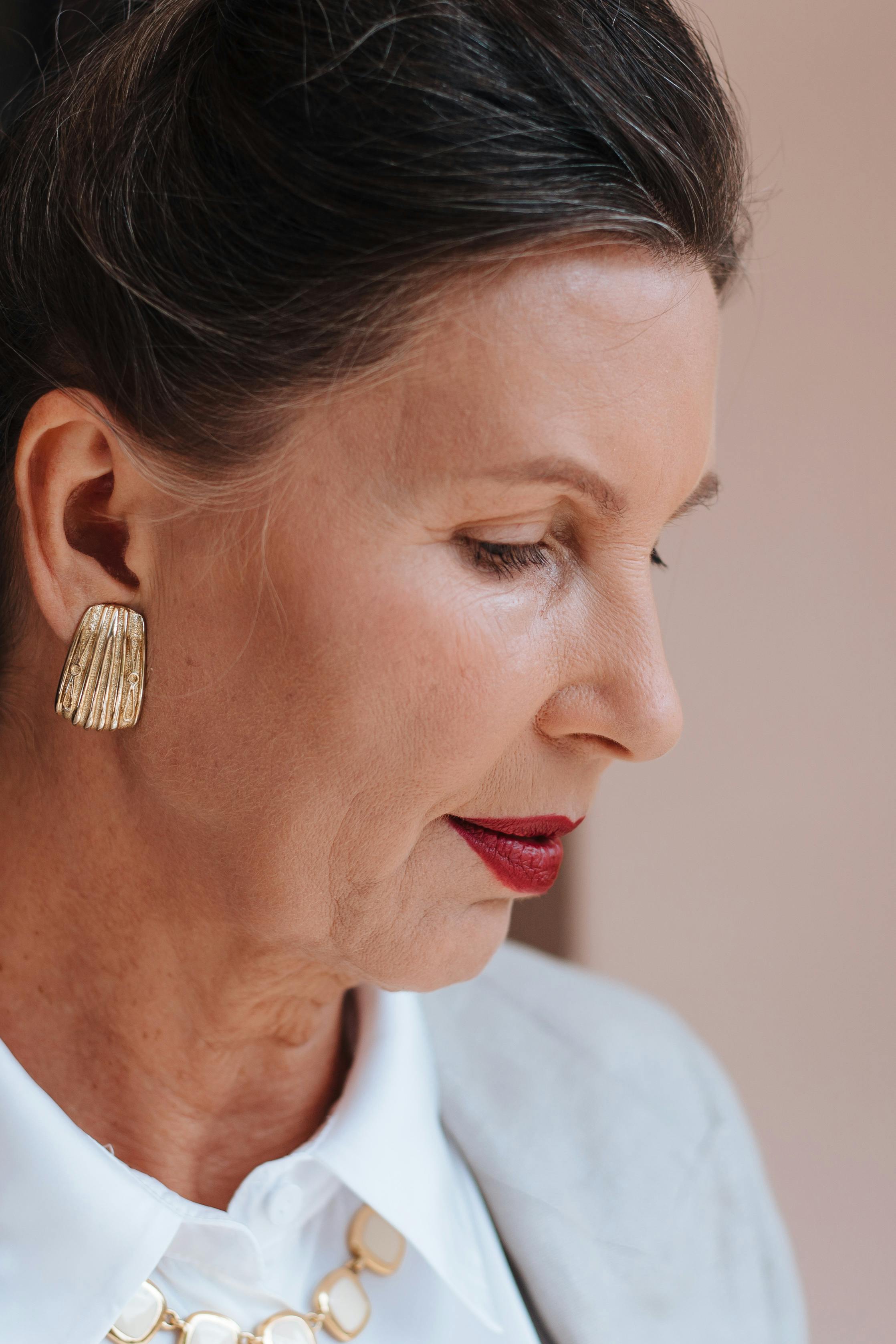 Profile view of a mature woman in elegant attire, showcasing gold earring and red lipstick.